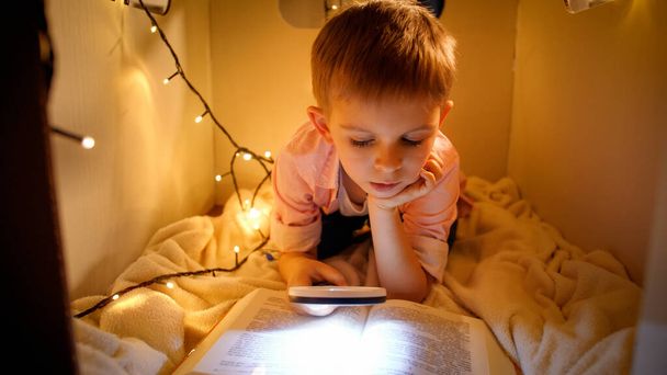 Porträt eines kleinen schlauen Jungen, der nachts ein großes Geschichtsbuch liest. Kind spielt in Spielzeugkartonhaus. Konzept der Kindererziehung und des Lesens im dunklen Raum. - Foto, Bild