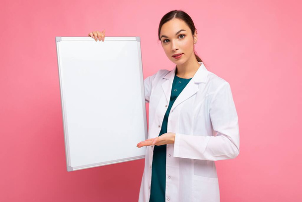 Portret atrakcyjnej młodej, pozytywnej brunetki ubranej w medyczny biały płaszcz z białą tablicą magnetyczną do makiety odizolowanej na różowym tle z przestrzenią do kopiowania - Zdjęcie, obraz
