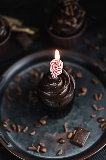 Several muffins or cupcakes with chocolate shaped cream at black table. Festive candle burns on a chocolate cake - Photo, Image