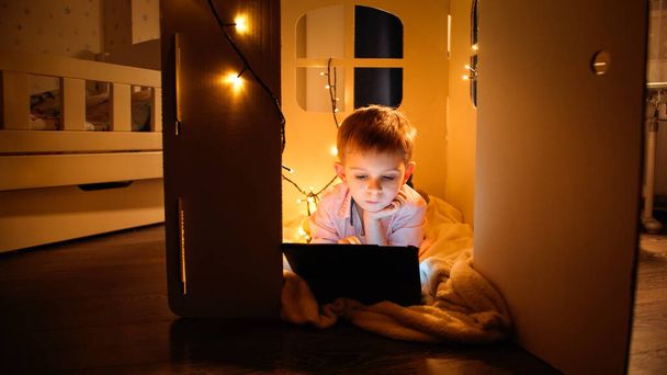小さなおもちゃの家に寝そべっている小さな賢い少年と夜にタブレットPCを争う。夜間の児童教育・学習の概念 - 写真・画像