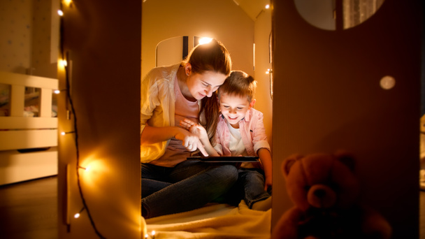 若い母親がゲームをプレイし、寝る前に夜にタブレットコンピュータを使用して幸せな笑顔の男の子。夜間に一緒に時間を持つ子供の教育と家族の概念 - 写真・画像