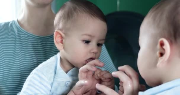 Söpö seitsemän kuukauden ikäinen vastasyntynyt vauva leikkii peilillä kotona äitinsä sylissä, näkee hänen heijastuksensa ja lyö kämmenensä peiliin. - Materiaali, video