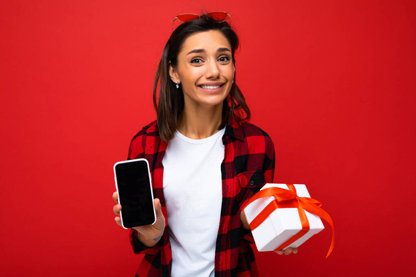 Belle jeune femme brune heureuse isolée sur un mur de fond rouge portant un t-shirt décontracté blanc et une chemise rouge et noire tenant une boîte cadeau blanche avec ruban rouge et téléphone portable avec écran vide - Photo, image