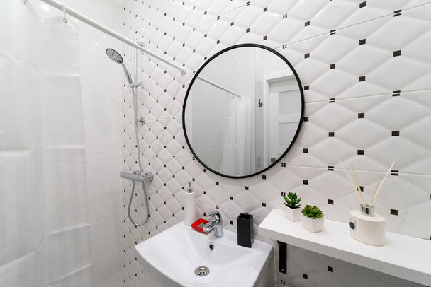 Helles, modernes Badezimmer. Ein Teil des Raumes mit einem weißen Waschbecken mit Wasserhahn, Regal, Dusche und einem runden Spiegel an der weiß-schwarzen Fliesenwand, der die Tür und einen weißen Vorhang reflektiert - Foto, Bild