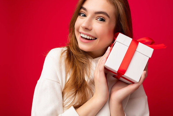 közelkép portré fénykép vonzó elragadó mosolygós szőke fiatal nő elszigetelt felett piros háttér fal visel alkalmi fehér kapucnis gazdaság fehér ajándék doboz piros szalaggal, és nézte - Fotó, kép