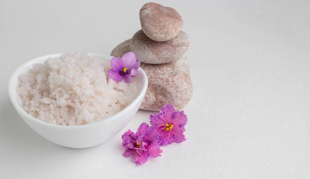 Розовая грубая соль в белой миске стоит на белом фоне рядом с розовыми камнями и розовыми фиолетовыми цветами. - Фото, изображение