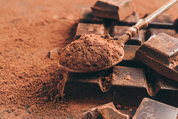 Braunes Kakaopulver im Löffel und gehackte Schokoladenwürfel auf dunklem Hintergrund. - Foto, Bild