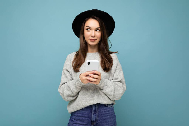 Beautiful young brunette woman thinking wearing black hat and grey sweater holding smartphone looking to the side texting isolated on background - Photo, Image