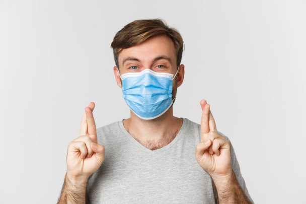 Concept of pandemic, covid-19 and social-distancing. Hopeful worried guy in medical mask, cross fingers for good luck and making wish, standing over white background - Photo, Image