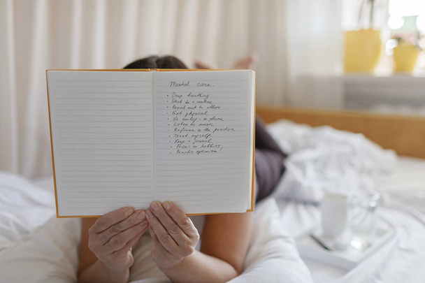 Mental health care, positive thinking and wellness concept. Attractive woman lying in bed, practicing stress relief challenge with journal about feeling, glass of water and gratitude list - Photo, Image