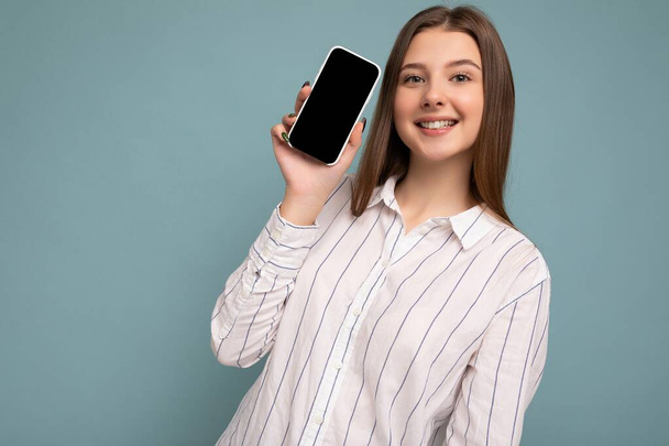 Photo of beautiful smiling young woman good looking wearing casual stylish outfit standing isolated on background with copy space holding smartphone showing phone in hand with empty screen display for - Foto, imagen