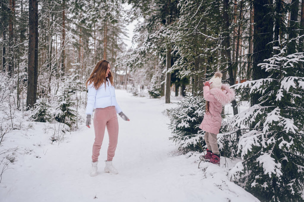 Mãe feliz e pequena menina bonito em roupas quentes rosa andando jogando luta bola de neve se divertindo na floresta de coníferas de inverno branco nevado com madeiras de abeto ao ar livre. Desporto familiar atividades de férias - Foto, Imagem