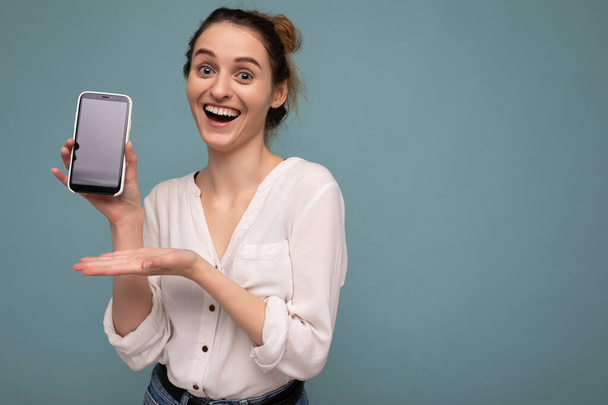 Положительная счастливая молодая блондинка кудрявая женщина в случайной белой рубашке и изолированная на голубом фоне стена держит телефон и показывает смартфон с пустым экраном для макета, глядя в камеру - Фото, изображение