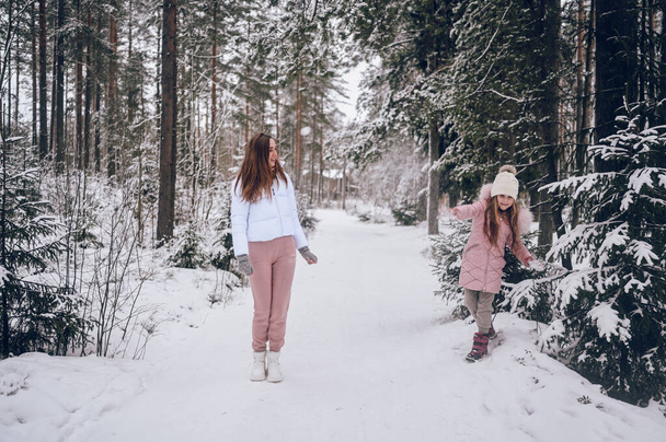 Madre feliz y niña linda en ropa interior de color rosa cálido caminar jugando lucha bola de nieve divertirse en el bosque de coníferas de invierno blanco nevado con maderas de abeto al aire libre. Deportes de familia actividades de vacaciones - Foto, imagen