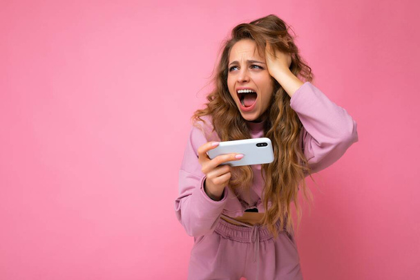 Photo of attractive crazy amazed surprised young woman wearing casual stylish clothes standing isolated over background with copy space holding and using mobile phone looking to the side and shouting - Φωτογραφία, εικόνα