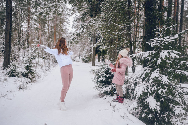 Madre feliz y niña linda en ropa interior de color rosa cálido caminar jugando lucha bola de nieve divertirse en el bosque de coníferas de invierno blanco nevado con maderas de abeto al aire libre. Deportes de familia actividades de vacaciones - Foto, Imagen