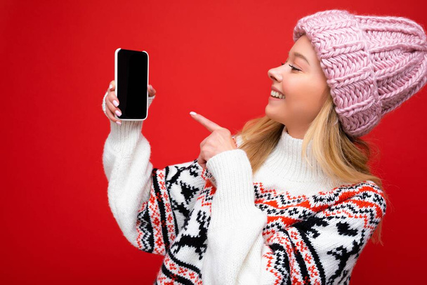 Foto van aantrekkelijke glimlachende jonge blonde vrouw met warme gebreide hoed en de winter warme trui staan geïsoleerd over rode achtergrond toont smartphone met leeg scherm voor mock up kijken naar apparaat - Foto, afbeelding