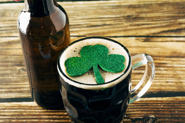 Zbliżenie pianki ciemnego piwa z błyszczącym liściem koniczyny, serwowane w kubku na drewnianym stole. Koncepcja St. Patrick i Stout - Zdjęcie, obraz