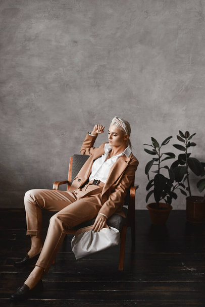 Young model woman in a trendy beige suit posing in a vintage armchair in minimalist interior - 写真・画像