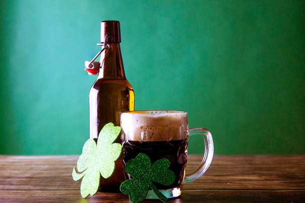 A mug of dark beer with its open bottle on a green background and a wooden table, next to them two shiny leaves of green clovers symbol of good luck. St. Patrick and stout concept - Photo, image