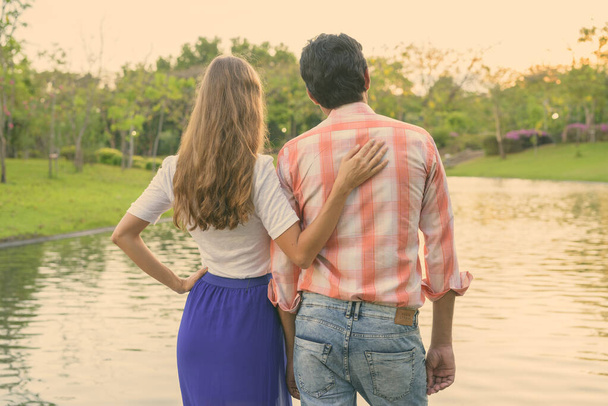 Обратный вид на многонациональную пару, стоящую и наслаждающуюся живописным видом на озеро в мирном зеленом парке вместе - Фото, изображение