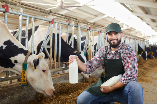 Smiling man farmer in uniform and rubber boots sitting and feeding cows in stall with hay - Φωτογραφία, εικόνα