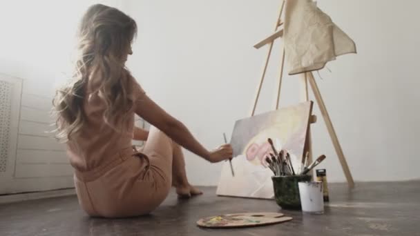 Achteraanzicht van vrouwelijke kunstenaar zit op de vloer en schildert beeld in haar atelier, het creëren van een prachtig beeld, schilderen met kleurrijke oliën. - Video