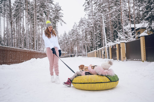 Happy mother and little cute girl in pink warm outwear walking having fun rides inflatable snow tube with red shiba inu dog in snowy white winter countryside outdoors. Family sport vacation activities - Photo, Image