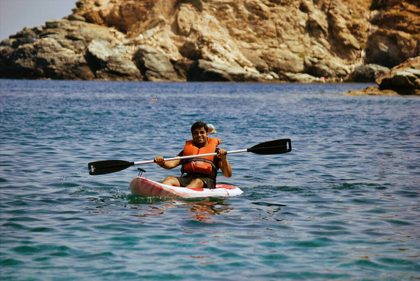 Kreta or Crete, Greece - Ένας άντρας με σωσίβιο να επιπλέει πάνω σε καγιάκ σε μια θάλασσα με φόντο ένα θολό βραχώδες βουνό. Θαλάσσια σπορ καγιάκ κατά τη διάρκεια του καλοκαιριού - Φωτογραφία, εικόνα