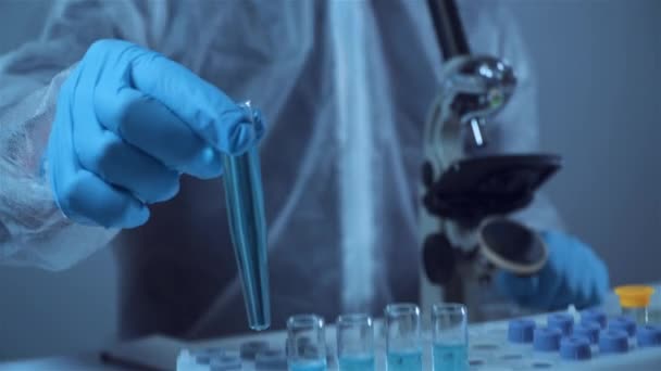 Koruyucu tulum giyen bilim adamı ya da doktor mavi reaktörlü test tüpü, kimyasal sıvı, mikroskop, laboratuvar biyokimyası içeren laboratuvar camları alıyor. Tıp ve araştırma konsepti - Video, Çekim
