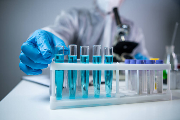 Wetenschapper of arts in beschermende overalls, neemt reageerbuis met blauw reagens, laboratorium glaswerk met chemische vloeistof, microscoop, laboratorium biochemie. Geneeskunde en onderzoek - Foto, afbeelding