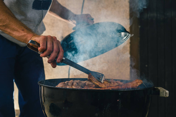 Man opening a smoker barbecue or a charcoal grill oven, to turn kebabs or spare ribs grilling on heat. Smoke visible rising from grill - Photo, Image