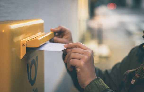 Mains d'une jeune femme blanche caucasienne vue insérer une enveloppe postale ou une lettre dans une boîte aux lettres jaune dans la soirée. Courrier tardif avec une enveloppe blanche. - Photo, image