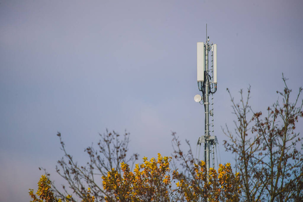 GSM антенны на вершине башни с облаками и небом на заднем плане и деревьями на переднем плане. Мобильная вышка 5G 4G и 3G LTE для антенны. Телекоммуникационная мачта. - Фото, изображение