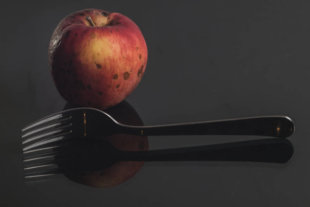 A natural looking red apple on a shiny reflective surface accompanied by a fork. Concept of healthy living and eating fresh natural produced fruit. - Photo, Image