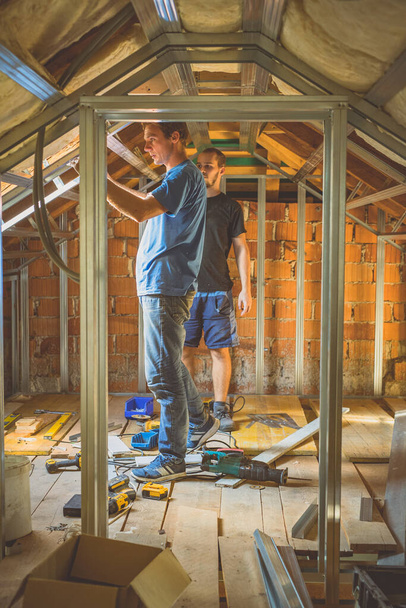 Dos hombres renovando su casa, construyendo una construcción de metal para paneles de yeso en un ático o loft. Feliz hombre caucásico renovando una casa con la ayuda de un amigo. - Foto, Imagen