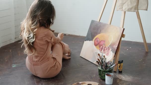 Achteraanzicht van vrouwelijke kunstenaar zit op de vloer en schildert beeld in haar atelier, het creëren van een prachtig beeld, schilderen met kleurrijke oliën. - Video