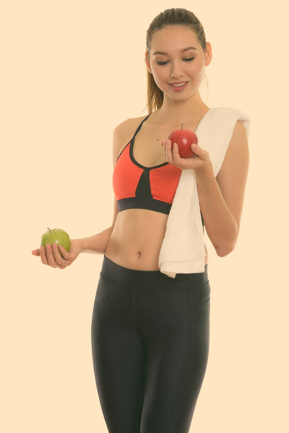 Studio shot of young happy beautiful Asian woman smiling and standing while looking at red apple and holding green apple - Photo, image