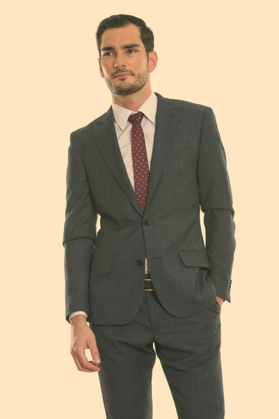Студийный снимок молодого привлекательного бизнесмена, стоящего, думая рукой о кармане - Фото, изображение