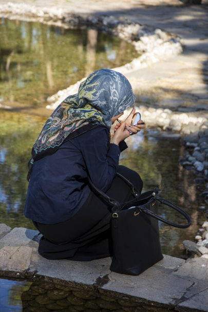 Muslim girl putting makeup in the park. Female model holding mirror and putting lipstic while she is in the nature. - Fotó, kép