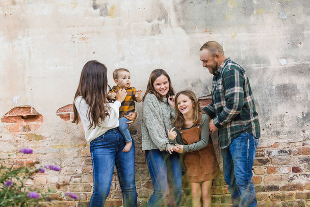 Μια πενταμελής οικογένεια με δύο κορίτσια και ένα αγοράκι να στέκονται δίπλα σε έναν τοίχο από τούβλα και να γελάνε. - Φωτογραφία, εικόνα