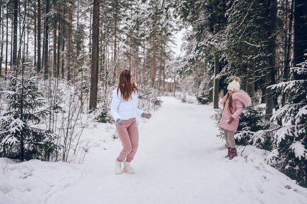 Happy mother and little cute girl in pink warm outwear walking playing snowball fight having fun in snowy white winter coniferous forest with spruce woods outdoors. Family sport vacation activities - Photo, Image