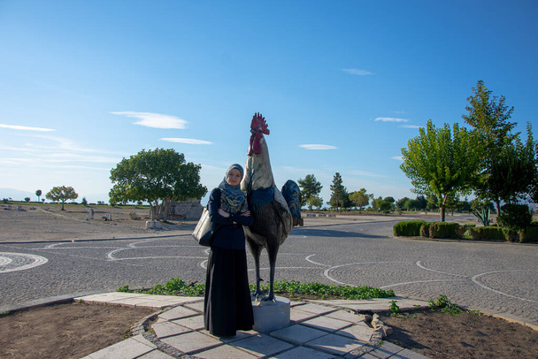Muslim girl standing next to the rooster sculpture at Pamukkale park in Denizli city. Travel and visit famous touristic destination in Turkey - Photo, Image