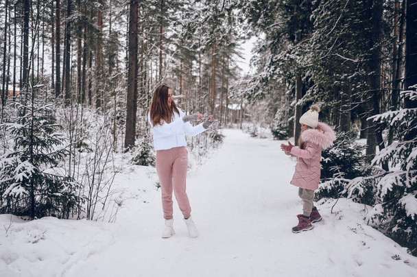 Happy mother and little cute girl in pink warm outwear walking playing snowball fight having fun in snowy white winter coniferous forest with spruce woods outdoors. Family sport vacation activities - Photo, Image