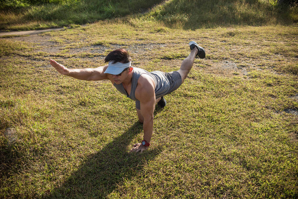 Un hombre asiático en forma y joven realiza perros pájaro en la hierba de un campo abierto. Parte inferior de la espalda y entrenamiento ab. - Foto, imagen