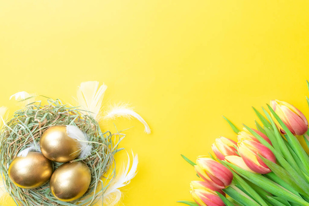 Egg color. Happy Easter decoration: golden colour eggs in basket with spring tulips, white feathers on pastel yellow background. Foil minimalist egg design, modern top view template - Photo, Image