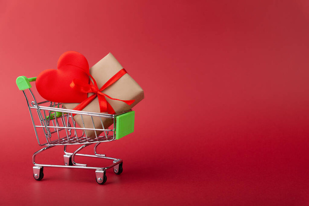 Red heart, gift box with red ribbon inside mini grocery cart on colored background - Foto, immagini