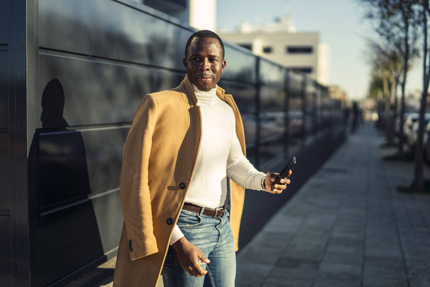 Μια ρηχή φωτογραφία ενός όμορφου, χαμογελαστού Αφρικανού με παλτό και τηλέφωνο στα χέρια. - Φωτογραφία, εικόνα