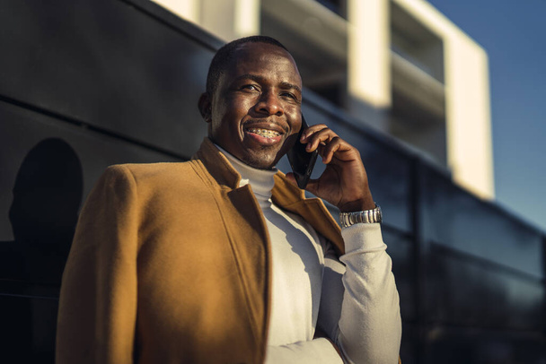 A shallow focus shot of a stylish handsome smiling African male in a coat talking by phone - Photo, image