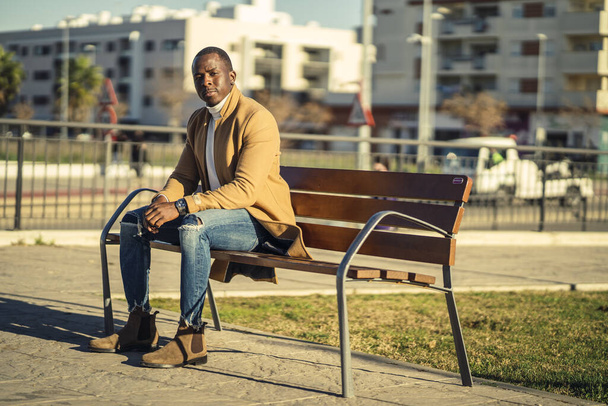 A closeup shot of a stylish handsome African male in a coat sitting on a bench and looking at camera - Фото, изображение